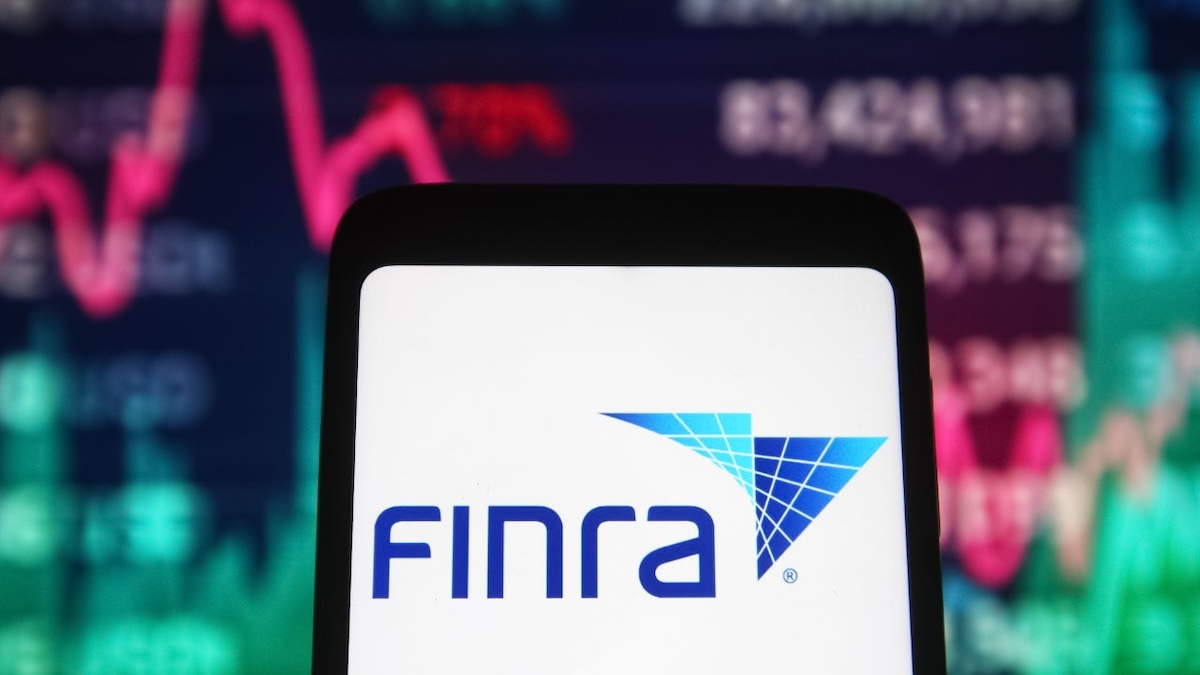 A mobile device displaying the FINRA (Financial Industry Regulatory Authority) logo on its screen. In the background, there is a blurred view of a stock market chart with red and green line graphs and numerical data, indicating financial and stock trading activity. The scene highlights the connection between FINRA and financial markets.