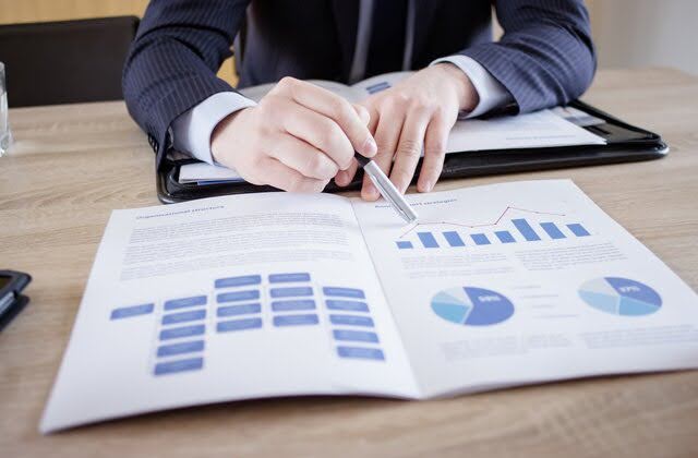 A financial advisor in a suit is pointing to a document with charts and graphs during a meeting. The document includes a flowchart on one page and various financial graphs and data on another.