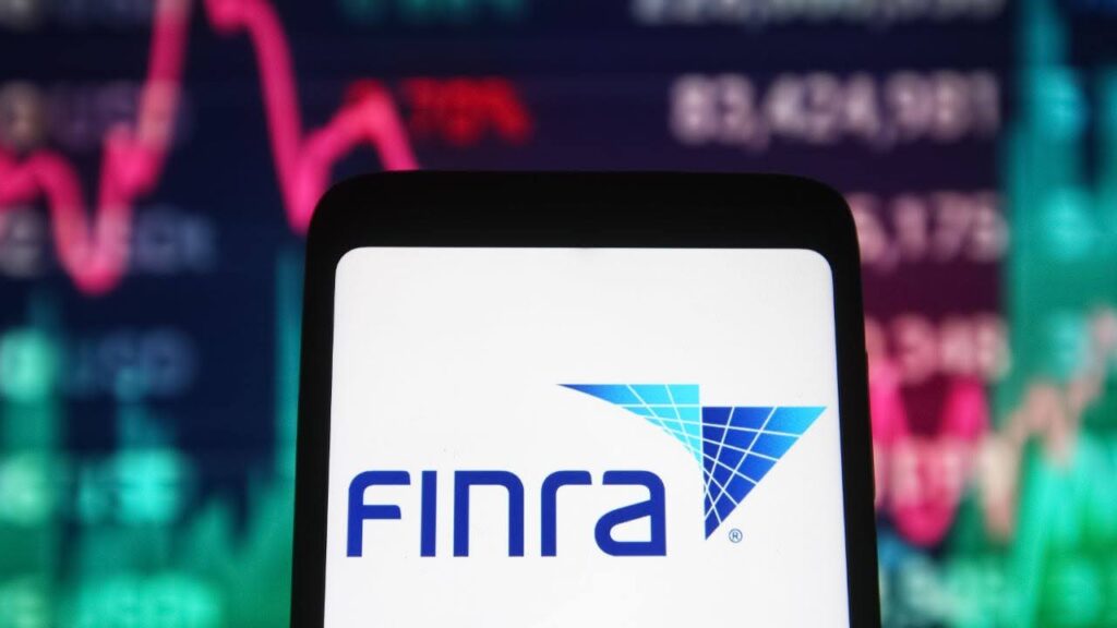 The image shows a mobile device displaying the FINRA (Financial Industry Regulatory Authority) logo on its screen. In the background, there is a blurred view of a stock market chart with red and green line graphs and numerical data, indicating financial and stock trading activity. The scene highlights the connection between FINRA and financial markets.