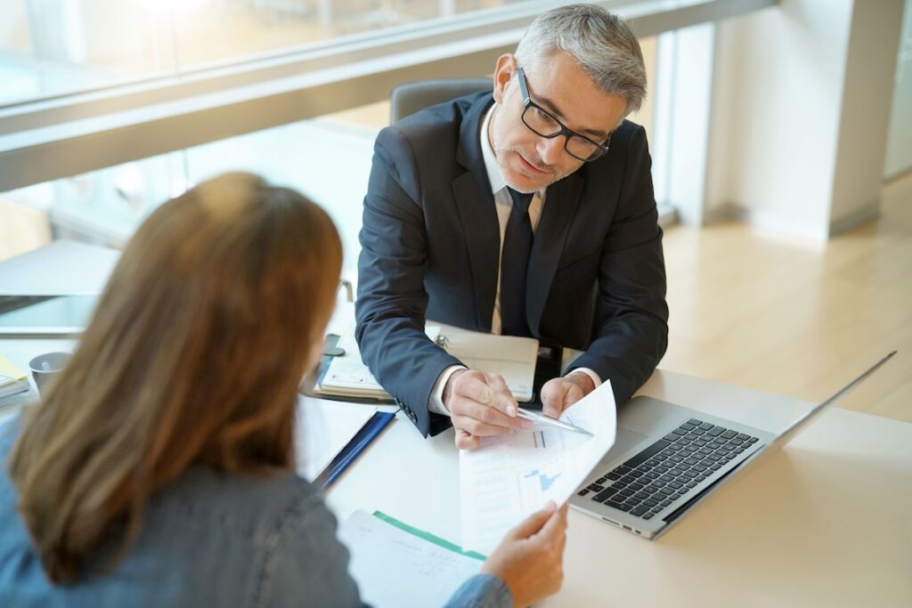 A male legal professional wearing glasses, sitting at a desk and reviewing documents with a female client in an office setting.