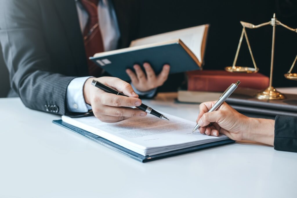 Two individuals signing legal documents at a desk, with law books and a scale of justice visible in the background.