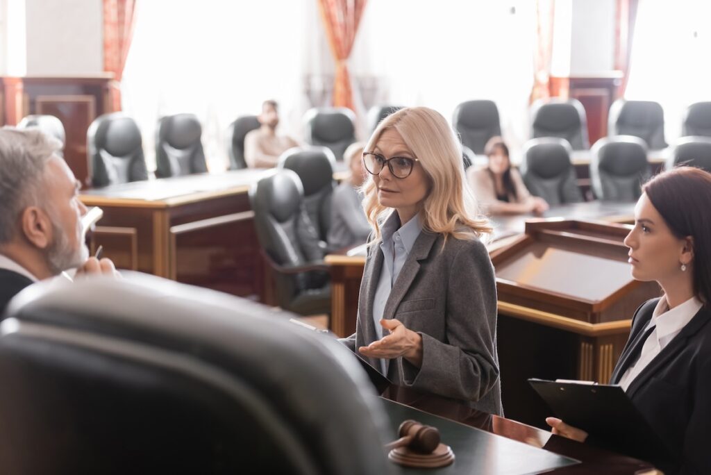 A female lawyer speaks to a judge in a courtroom, with attendees seated in the background and another lawyer standing beside her holding documents.