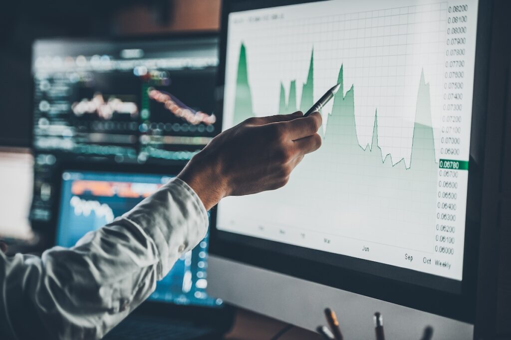 A person pointing at a graph on a computer screen, analyzing market data with various charts displayed in the background, representing financial analysis and stock market trends.