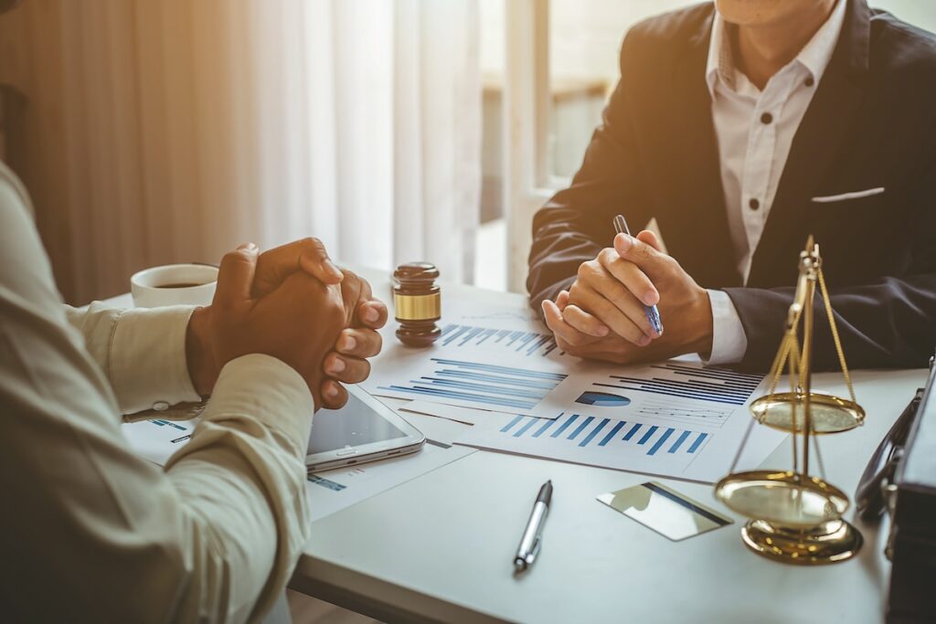 Two professionals sitting at a table during a business meeting, with charts, scales of justice, and a gavel visible on the desk.