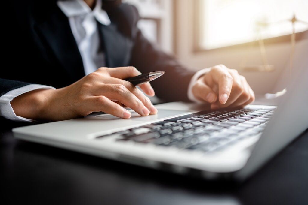 Close-up of a businessperson's hands using a laptop while holding a pen, suggesting work or research in a professional setting.