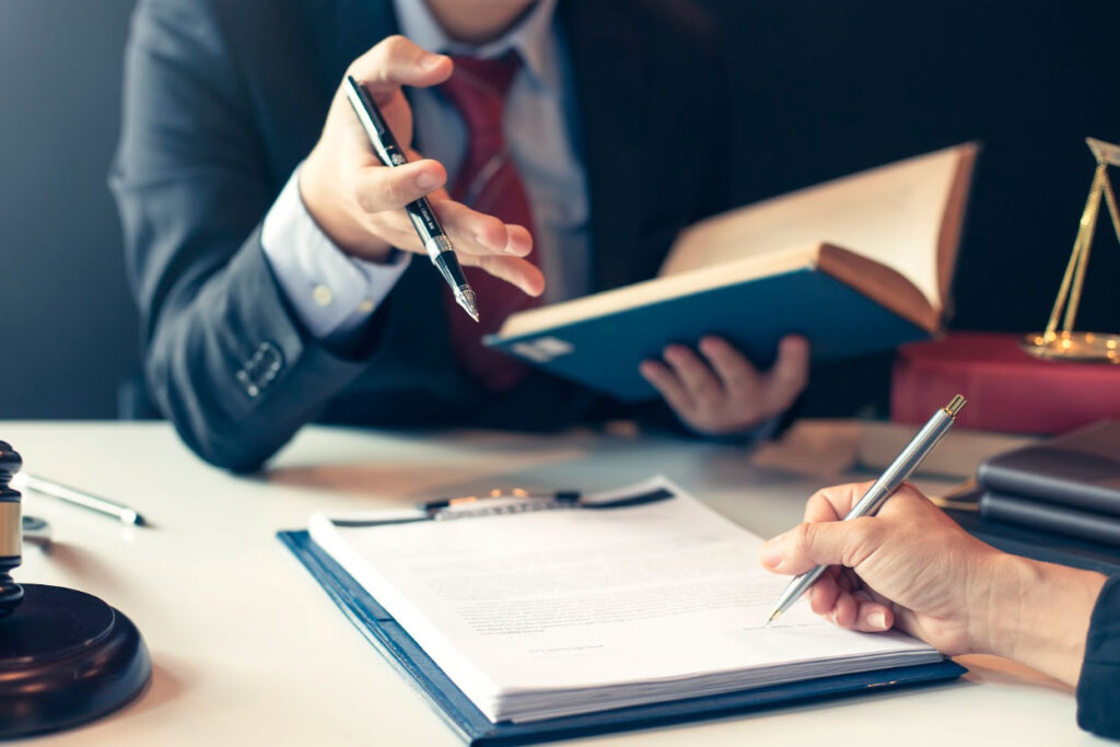 Two professionals discussing legal documents, with one holding a pen and the other holding a book, alongside a gavel and scales of justice on the table.