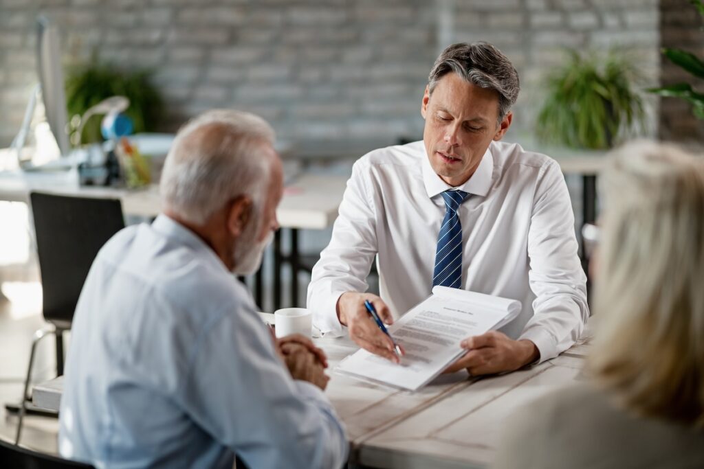 A lawyer in a white shirt and blue tie is seated at a table, explaining legal documents to an older couple. The lawyer is pointing at the paperwork while the clients listen attentively in a professional office setting