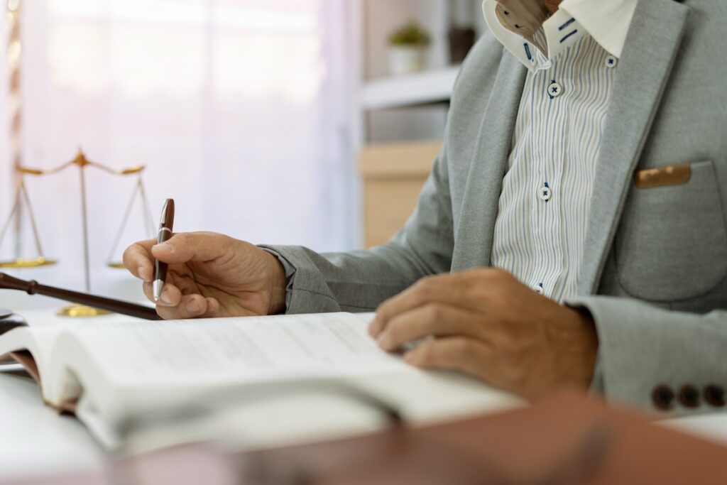  A lawyer in a suit is writing in an open notebook at a desk with golden scales of justice and legal documents nearby. Several law books are stacked in the background, highlighting the professional legal environment.