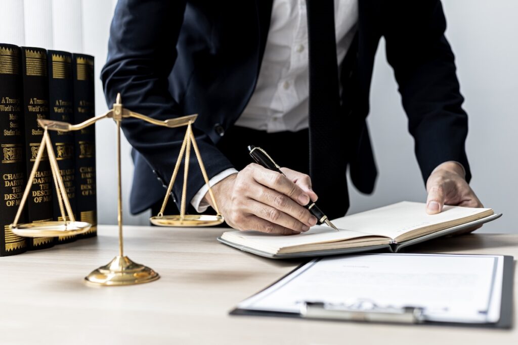 A lawyer in a suit is writing in a notebook at a desk, with a set of golden scales of justice and several legal books in the background.