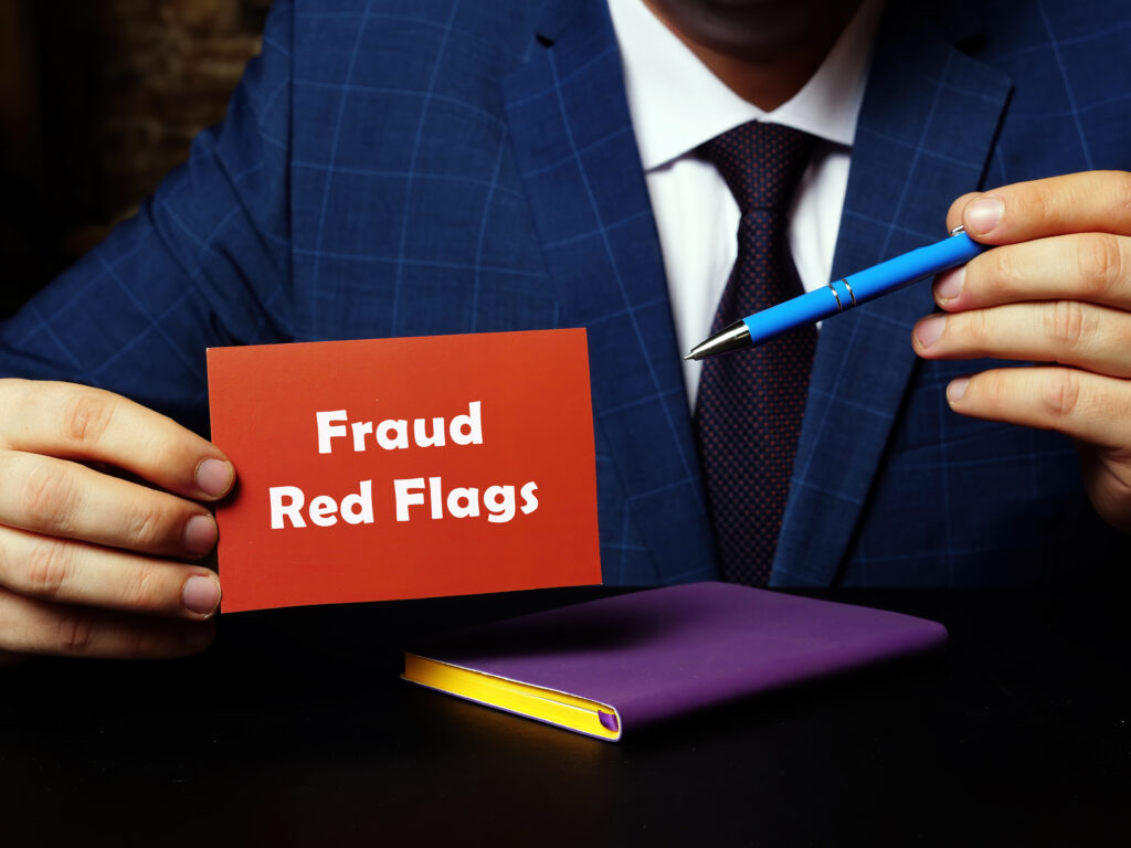 A person in a blue suit holding a red card with the text "Fraud Red Flags" written on it, while pointing at the card with a blue pen. A purple notebook is visible on the desk below.