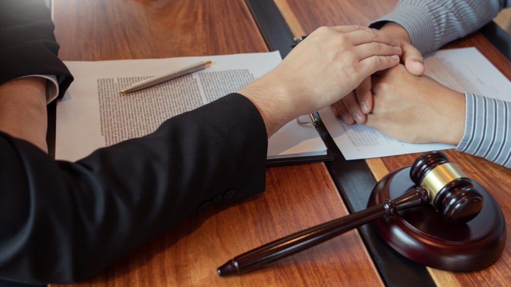 Close-up of a lawyer offering comfort by holding the hands of a client across a desk, with legal documents, a pen and a gavel visible on the table.