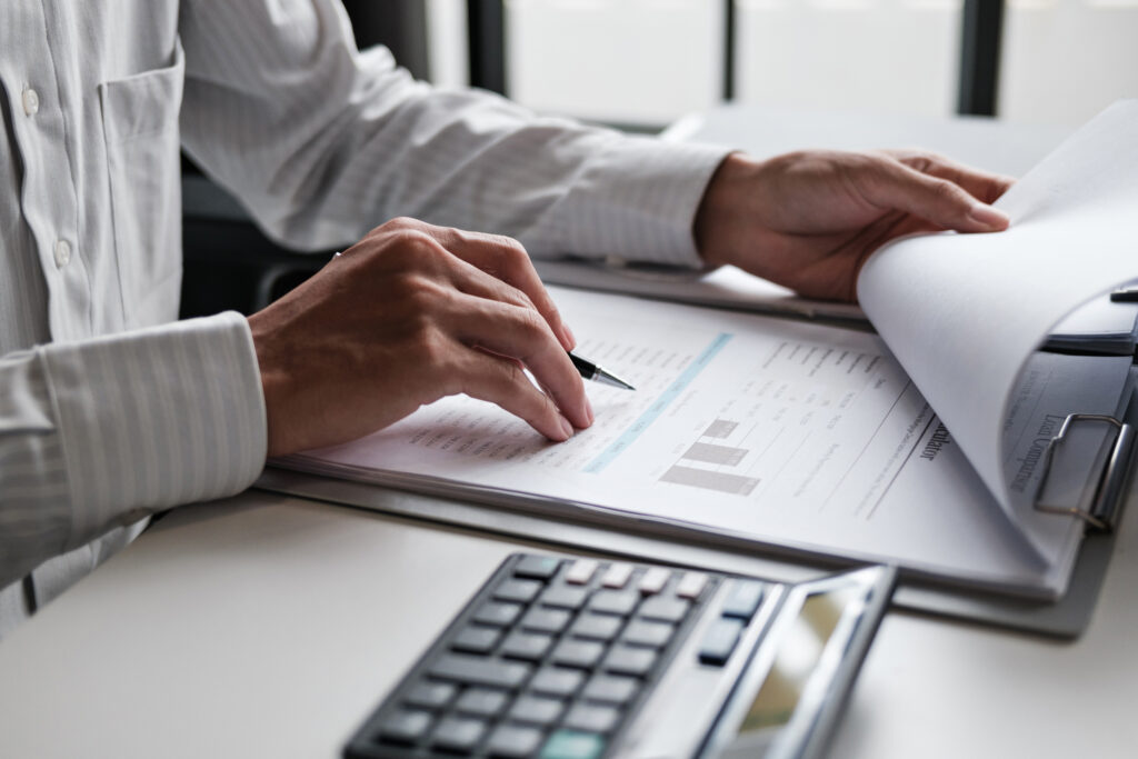 Close-up of a person reviewing financial documents with charts and graphs, using a pen and calculator at a desk.